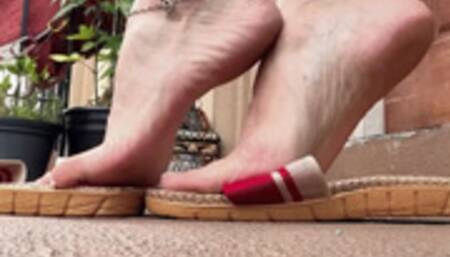 Slippers dangle, arches, and soles show, below my chair pov, long sexy red dress, white toes